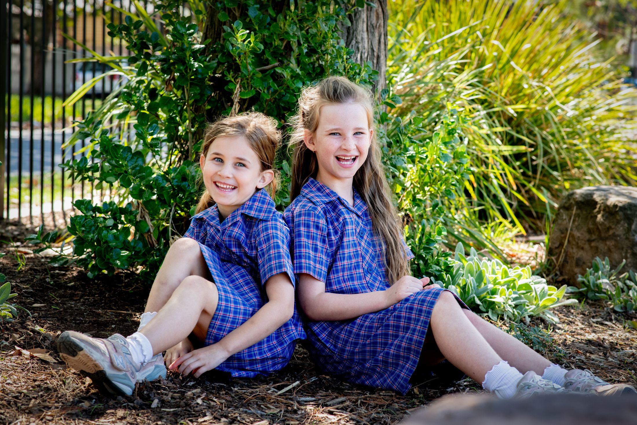 Two students in school uniforms laughing together, sitting on the ground in a garden